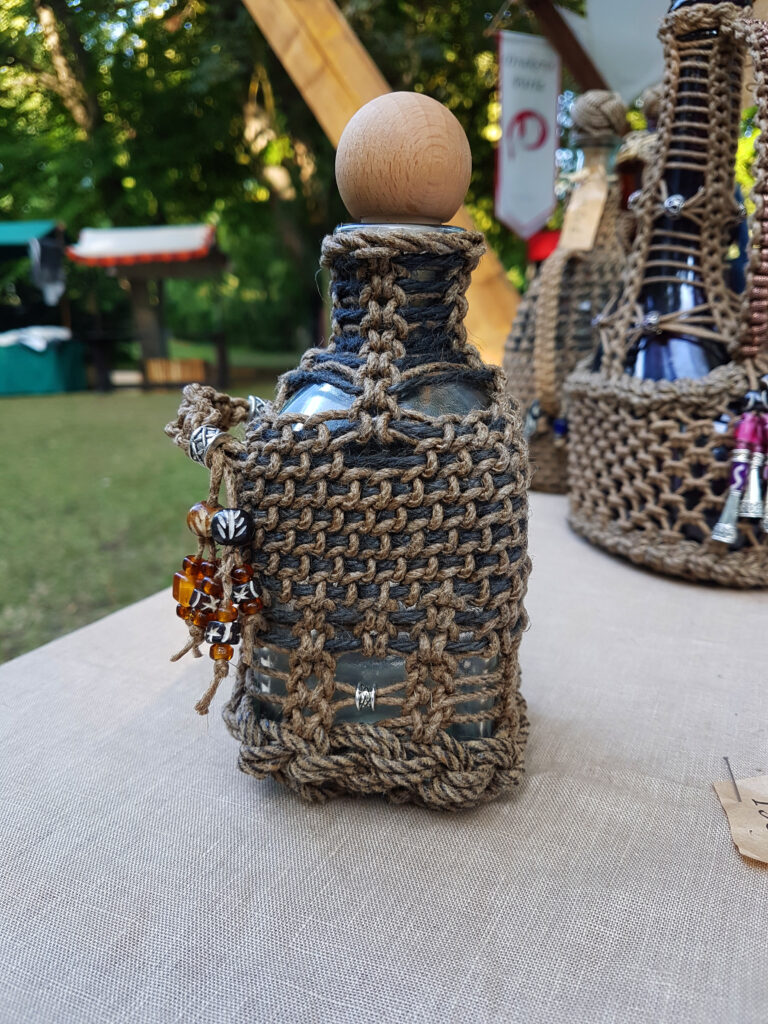 Mindre inknuten glasflaska med träkork, omsluten av handknutet hampenät och dekorerad med bärnstensfärgade pärlor.