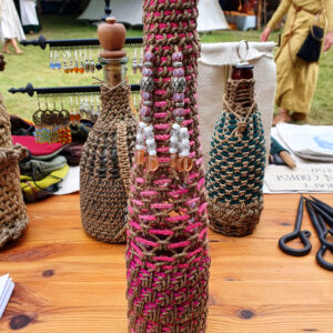 Inknuten glasflaska i grovt hamparep med infällda rosa snören samt dekorationer av glas- och metallpärlor, fotograferad på medeltidsmarknad.